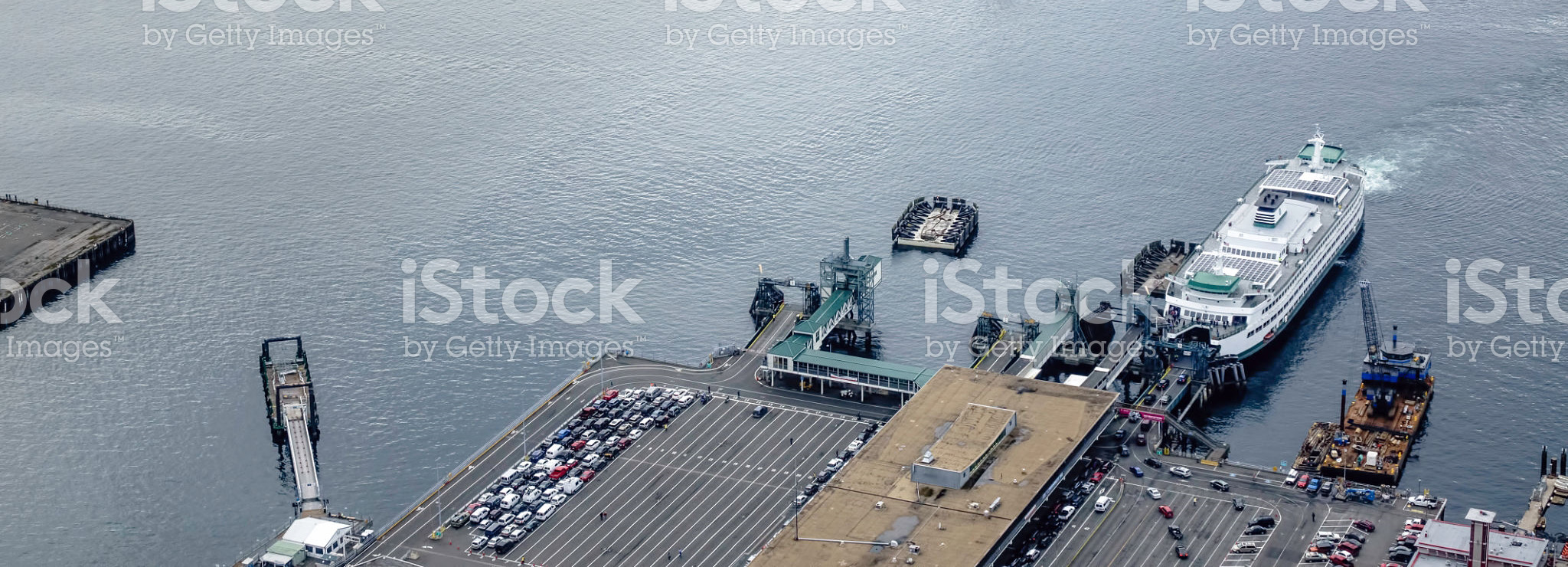 Aerial view of a ferry terminal in Seattle, with one ferry docked and another approaching lines of parked vehicles in wait (pedestrians are too small to be identified)